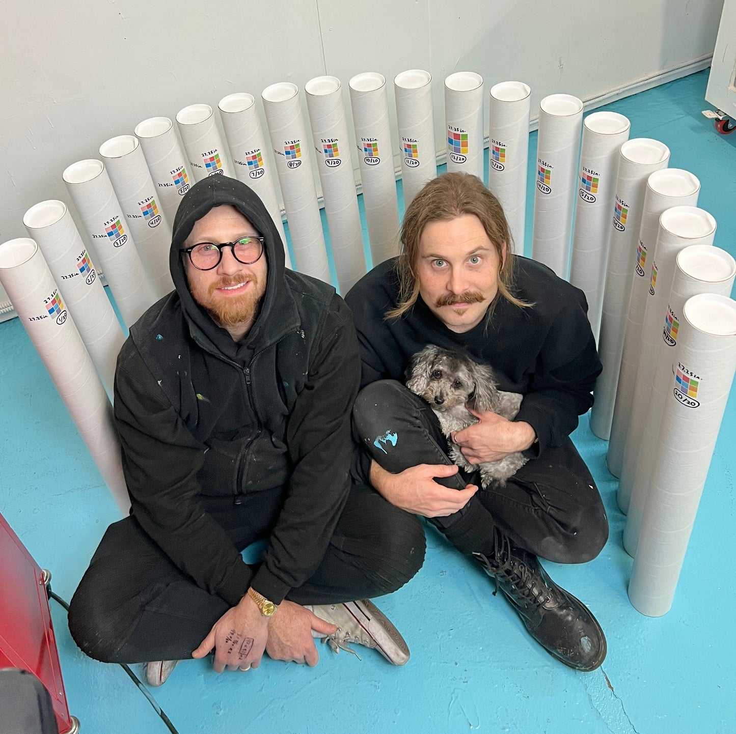 Two people sitting on a blue floor with rolls of paper and a small dog.
